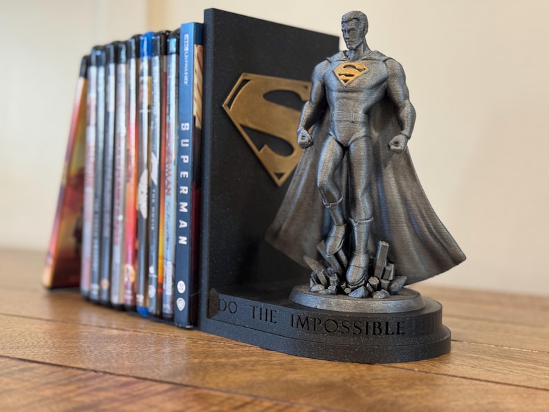 May include: A Superman-themed bookend set. The set includes a black bookend with a gold Superman logo and a gray Superman figure. The base of the figure reads "DO THE IMPOSSIBLE". Blu-ray discs are visible.
