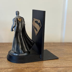 May include: A black and gray Superman bookend set. One bookend features a Superman figure with a cape and the Superman logo on the back. The other bookend is a black rectangular shape with the Superman logo.