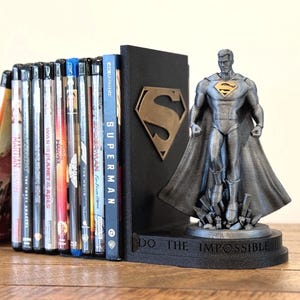 May include: A black Superman bookend with a bronze-colored "S" logo and a Superman figurine. The bookend supports a collection of DVDs and Blu-ray discs. The base of the bookend reads "DO THE IMPOSSIBLE".