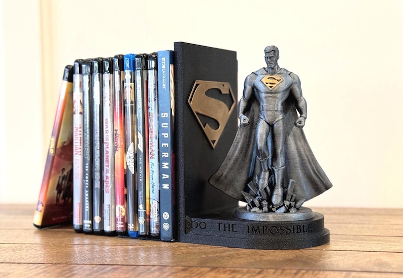 May include: A black Superman bookend with a bronze-colored "S" logo and a Superman figurine. The bookend supports a collection of DVDs and Blu-ray discs. The base of the bookend reads "DO THE IMPOSSIBLE".
