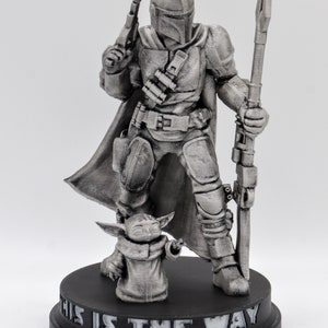 May include: A silver-colored figurine of a Mandalorian warrior holding a weapon and standing next to a small, silver-colored creature. The base of the figurine reads "This is the way".