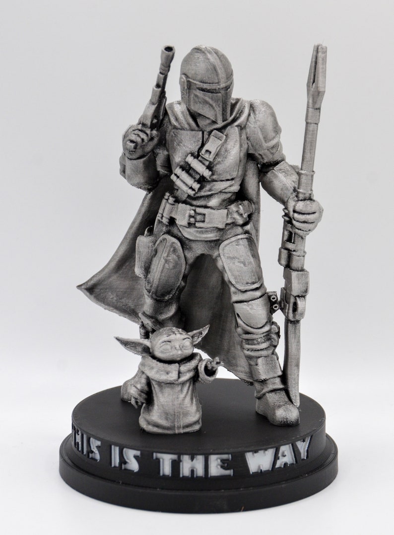May include: A silver-colored figurine of a Mandalorian warrior holding a weapon and standing next to a small, silver-colored creature. The base of the figurine reads "This is the way".