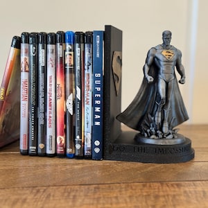 May include: A collection of Blu-ray movies and a DVD, held between a black Superman bookend and a Superman figurine. The bookend has a Superman logo and the figurine is on a base that reads "DO THE IMPOSSIBLE".