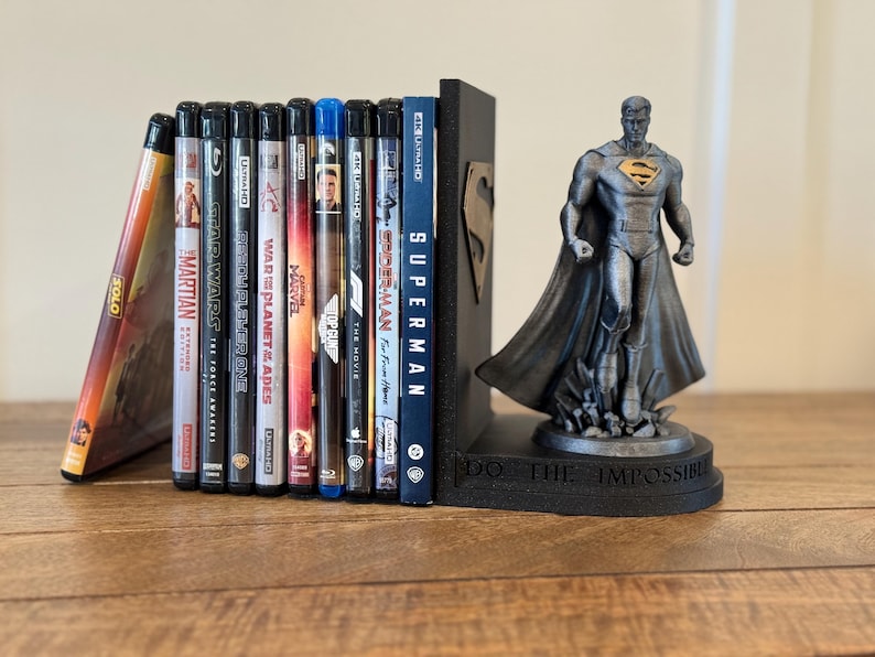 May include: A collection of Blu-ray movies and a DVD, held between a black Superman bookend and a Superman figurine. The bookend has a Superman logo and the figurine is on a base that reads "DO THE IMPOSSIBLE".