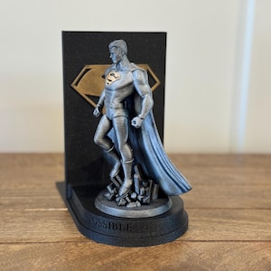 May include: A silver-colored Superman statue stands on a black base with the word "POSSIBLE" and a black background with a gold emblem. The statue is a detailed representation of the superhero.