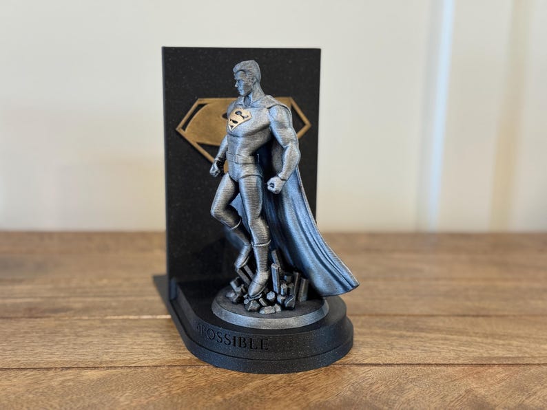May include: A silver-colored Superman statue stands on a black base with the word "POSSIBLE" and a black background with a gold emblem. The statue is a detailed representation of the superhero.