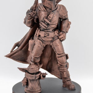 May include: A bronze-colored 3D printed figurine of a Mandalorian warrior standing with a blaster rifle in one hand and a cape flowing behind him. A smaller bronze-colored figurine of a Grogu character stands in front of the Mandalorian. The base of the figurine is black and has the words "This is the way" inscribed on it.