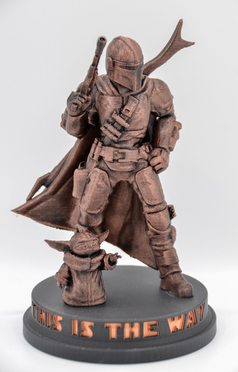 May include: A bronze-colored 3D printed figurine of a Mandalorian warrior standing with a blaster rifle in one hand and a cape flowing behind him. A smaller bronze-colored figurine of a Grogu character stands in front of the Mandalorian. The base of the figurine is black and has the words "This is the way" inscribed on it.