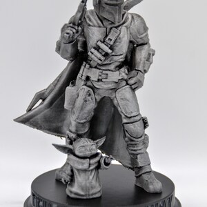 May include: A 3D printed figurine of a Mandalorian character from the Star Wars franchise, standing with a small, grey alien figure. The Mandalorian is wearing a helmet and armor, holding a blaster rifle. The base of the figurine is black and reads "This is the way".
