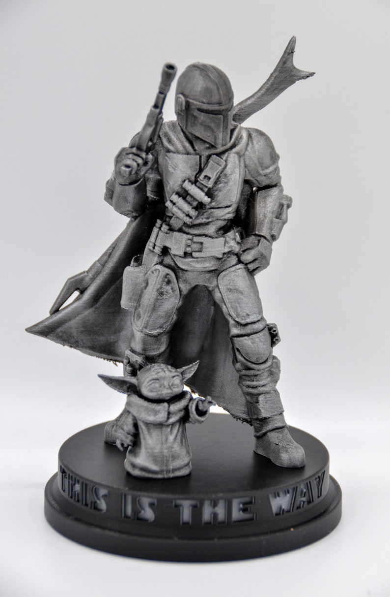 May include: A 3D printed figurine of a Mandalorian character from the Star Wars franchise, standing with a small, grey alien figure. The Mandalorian is wearing a helmet and armor, holding a blaster rifle. The base of the figurine is black and reads "This is the way".