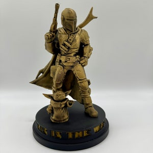 May include: A gold-colored 3D printed figurine of a Mandalorian character from Star Wars, standing with a blaster rifle and a cape. The character is standing on a black base with the words "This is the Way" inscribed on it. A smaller gold-colored figurine of the character "The Child" is standing at the Mandalorian's feet.