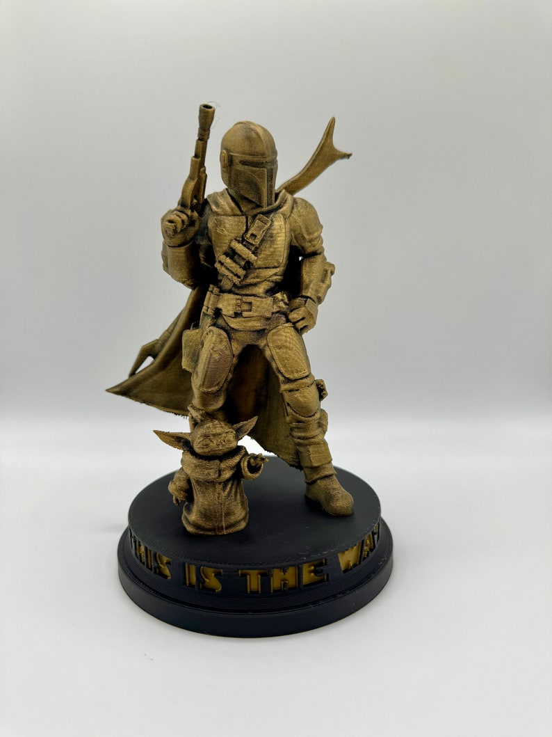 May include: A gold-colored 3D printed figurine of a Mandalorian character from Star Wars, standing with a blaster rifle and a cape. The character is standing on a black base with the words "This is the Way" inscribed on it. A smaller gold-colored figurine of the character "The Child" is standing at the Mandalorian's feet.