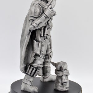 May include: A silver-colored figurine of a Mandalorian warrior with a cape and a blaster rifle, standing next to a small, silver-colored figurine of a baby alien. The base of the figurine is black and has the text "THIS IS THE WAY" printed on it.