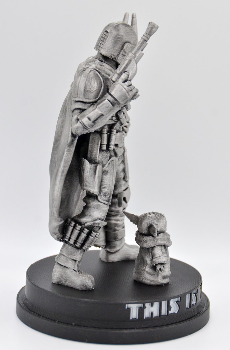 May include: A silver-colored figurine of a Mandalorian warrior with a cape and a blaster rifle, standing next to a small, silver-colored figurine of a baby alien. The base of the figurine is black and has the text "THIS IS THE WAY" printed on it.