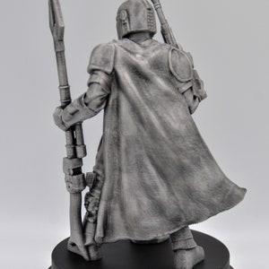 May include: A silver-colored figurine of a Mandalorian warrior holding a staff. The figure is wearing a cape and helmet and is standing on a black base.