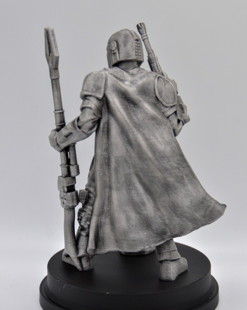 May include: A silver-colored figurine of a Mandalorian warrior holding a staff. The figure is wearing a cape and helmet and is standing on a black base.