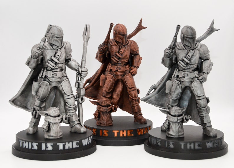 May include: Three 3D printed figurines of the Mandalorian character from the Star Wars series. The figures are painted in silver, copper, and bronze. Each figure is holding a weapon and has a small figure of Grogu, also known as Baby Yoda, standing at their feet. The base of each figure has the text "This is the Way" printed on it.