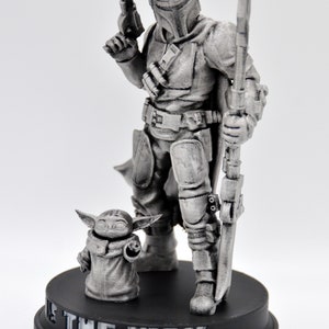 May include: A silver-colored figurine of a Mandalorian warrior standing with a rifle and a small, silver-colored creature standing in front of him. The base of the figurine is black and reads "This is the Way".