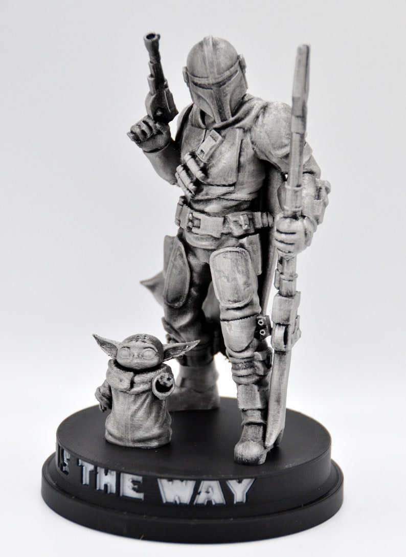May include: A silver-colored figurine of a Mandalorian warrior standing with a rifle and a small, silver-colored creature standing in front of him. The base of the figurine is black and reads "This is the Way".