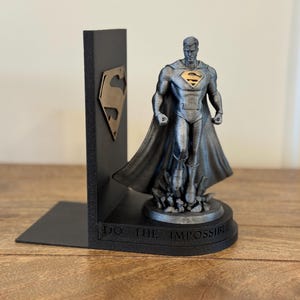 May include: A black Superman bookend with a gold Superman logo on the side. The bookend features a Superman statue with a cape and the words "DO THE IMPOSSIBLE" on the base. The statue is silver and gold.