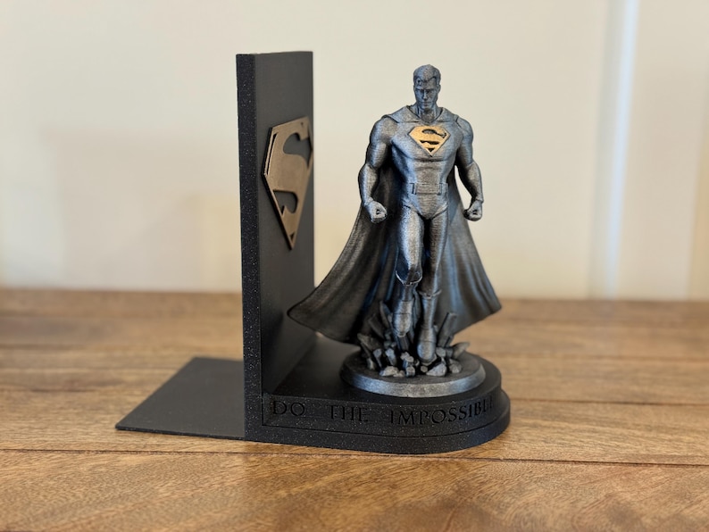 May include: A black Superman bookend with a gold Superman logo on the side. The bookend features a Superman statue with a cape and the words "DO THE IMPOSSIBLE" on the base. The statue is silver and gold.