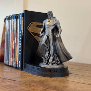 May include: A Superman-themed bookend set. The set includes a black bookend with a gold Superman logo and a silver Superman figure. The base of the bookend reads "DO THE IMPOSSIBLE". A collection of books is held in place.