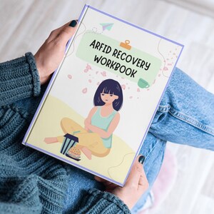 Pu&ograve; includere: Un quaderno di lavoro rosa e verde con il titolo "ARFID Recovery Workbook" e un'illustrazione di cartone animato di una persona seduta con una tazza di t&egrave;.