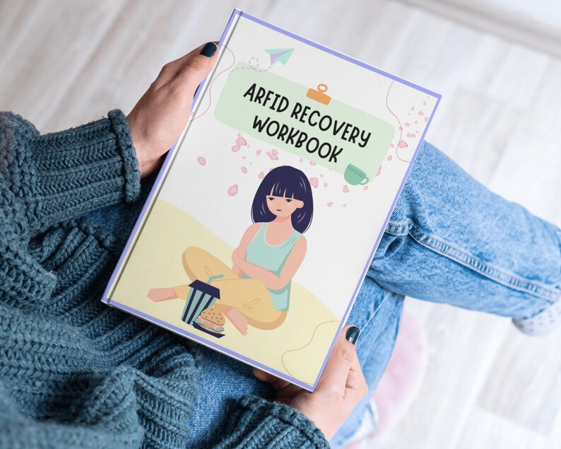 Pu&ograve; includere: Un quaderno di lavoro rosa e verde con il titolo "ARFID Recovery Workbook" e un'illustrazione di cartone animato di una persona seduta con una tazza di t&egrave;.
