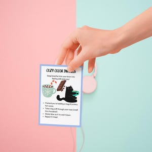 May include: A small card with the text "COZY COCOA INS..." is held by a hand. The card features illustrations of a mug of cocoa, a chocolate bar, and a black cat. The background is split between pink and light blue.
