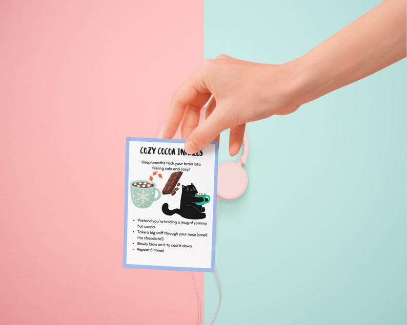 May include: A small card with the text "COZY COCOA INS..." is held by a hand. The card features illustrations of a mug of cocoa, a chocolate bar, and a black cat. The background is split between pink and light blue.