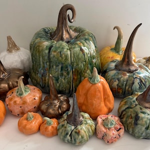 Op de afbeelding: Een verzameling keramische pompoenen in verschillende maten en kleuren, waaronder tinten groen, oranje, geel, wit en metallic brons. Elk stuk heeft een opvallende steel en een glanzende, getextureerde afwerking met zichtbare ribbels.