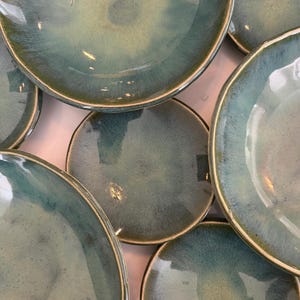May include: A collection of round ceramic plates and bowls featuring a reactive glaze in shades of teal, blue, and green. Each piece has a smooth, glossy finish with subtle variations in color intensity and a distinct, slightly darkened rim.