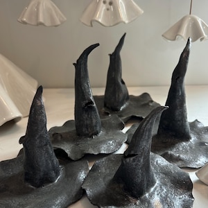 Puede incluir: Una colección de sombreros de bruja de cerámica hechos a mano en color gris oscuro, con un acabado texturizado. Los sombreros tienen forma puntiaguda y se asientan sobre un ala ancha. Encima, decoraciones blancas con forma de fantasma de cerámica.