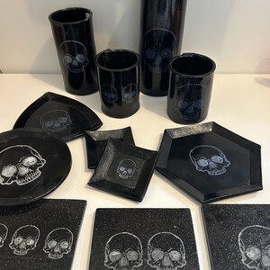 Op de afbeelding: Een verzameling zwart keramisch servies en drinkgerei met een schedelontwerp. De set bevat verschillende vormen van borden, onderzetters en cilindrische bekers en vazen. Het schedelontwerp is in een lichtere kleur, contrasterend met de zwarte achtergrond.