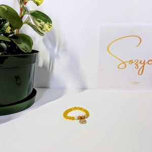 Könnte beinhalten: Ein gelbes Perlenarmband mit einem goldenen Anhänger liegt auf einer weißen Oberfläche. Das Armband hat gelbe, weiße und braune Perlen. Ein Blumentopf und ein Schild mit goldener Schrift befinden sich im Hintergrund. Auf dem Schild steht "Sozye".