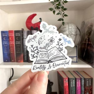 May include: A white sticker with a black and white illustration of an open book with bats and flowers flying out of it. The text "Reality Is Overrated" is written below the book.