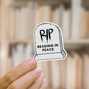 May include: A gray sticker in the shape of a tombstone with the letters "RIP" in black dripping font. Below the letters, the text reads "Reading in Peace". The sticker is held in a person's hand.