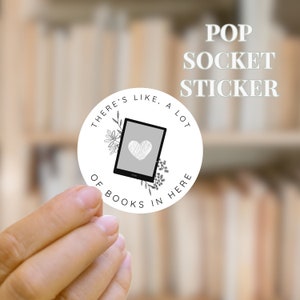May include: White pop socket sticker with the text "THERE'S LIKE, A LOT OF BOOKS IN HERE" surrounding a graphic of an e-reader with a heart. The words "POP SOCKET STICKER" are above the sticker.