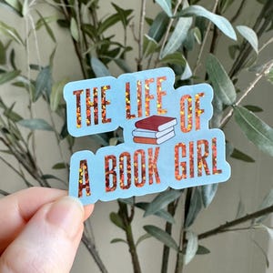 May include: A light blue sticker with the text "THE LIFE OF A BOOK GIRL" in orange glitter lettering. The sticker features a small illustration of three stacked books. The background includes green leaves.