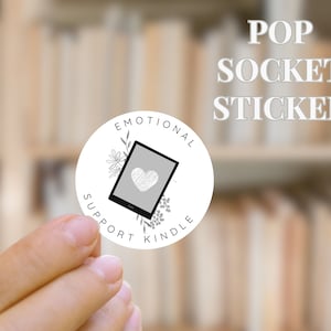May include: White round sticker with black text and a graphic of a Kindle e-reader with a heart on the screen. The text reads "Emotional Support Kindle". The sticker is being held by a hand. The text "POP SOCKET STICKER" is in the background.