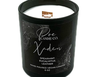 May include: A black glass candle jar with a white wax fill and a wooden wick. The label reads Poe Candle Co. Xaden, Spearmint Eucalyptus Leather, Hand Poured Vegan Candle, 6 oz. The label features a dark, abstract, textured background design.