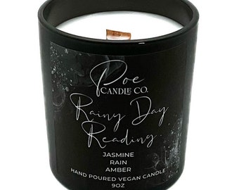May include: A black glass candle jar with a white wax interior and a wooden wick. The label features the text Poe Candle Co. Rainy Day Reading, Jasmine, Rain, Amber, Hand Poured Vegan Candle, and 9oz, set against a dark, abstract background.