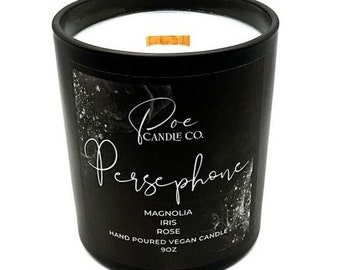 May include: A black glass candle jar with a white wax interior and a wooden wick. The label features the text Poe Candle Co., Persephone, Magnolia, Iris, Rose, Hand Poured Vegan Candle, and 9oz in white script and sans-serif lettering.