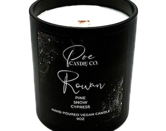 May include: A black cylindrical candle jar with a white wax top and a wooden wick. The label features the text Poe Candle Co., Rowan, Pine, Snow, Cypress, Hand Poured Vegan Candle, and 9oz in white lettering against a dark, textured background.