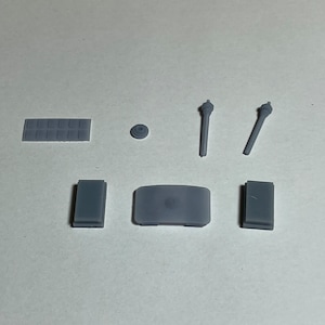 May include: An assortment of small, gray, plastic components, possibly for a model or a small device. The pieces include rectangular blocks, a small disc, two rod-like pieces, and a larger curved piece.