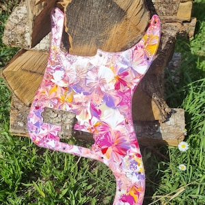 May include: A guitar pickguard with a vibrant floral design in shades of pink, purple, yellow, and white. The pickguard is set against a backdrop of wood and green grass, creating a colorful contrast. The floral pattern adds a touch of artistic flair.