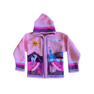 May include: A light pink knit zip-up hoodie featuring a hood with horizontal stripes in pink, purple, and blue. The front displays embroidered motifs including a yellow sun, a white cloud with rain, a cactus, a house, a lamb, a cat, and a bird.