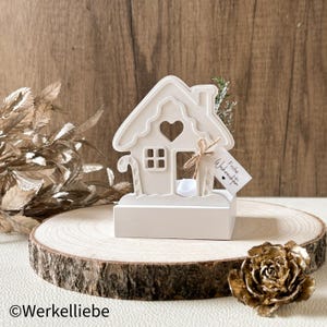 Könnte beinhalten: Ein cremefarbenes Lebkuchenhaus-Kerzenhalter mit herzförmigem Fenster und Zuckerstange. Ein Anhänger mit den Worten "Frohe Weihnachten" ist am Haus befestigt. Das Haus steht auf einer Holzscheibe.