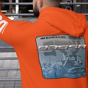 May include: Bright orange hoodie with white text reading "D355A" on the sleeve. The back features a graphic with the text "KOMATSU D355A-3 BULLDOZER" and an image of a bulldozer. The hoodie has a hood.