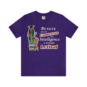 May include: Purple t-shirt with a graphic of a woman wearing a graduation cap and gown sitting on a stack of books. The text on the shirt reads "Beauty may be Dangerous but Intelligence is downright Lethal."